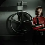 Tokyo, con la sua giacca rossa e nera, posa con le braccia conserte davanti alla porta di una cassaforte, un'immagine iconica de la casa di carta 4.