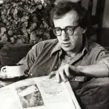 Woody Allen, con occhiali e camicia, legge un giornale con una tazza di caffè, forse pensando al suo prossimo Oscar.