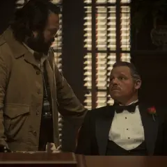 Un uomo con la barba e occhiali parla con un altro in smoking con una rosa. La scena suggerisce una recensione di un'offerta importante.