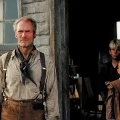 Un uomo anziano con bretelle e un fazzoletto al collo, sullo sfondo due bambini che osservano. La scena evoca il clima de "Gli spietati trama".