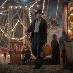 Un uomo con un cappello cammina in una fiera delle illusioni, con una ruota panoramica illuminata sullo sfondo. Le recensioni di Nightmare Alley descrivono questo mondo oscuro.