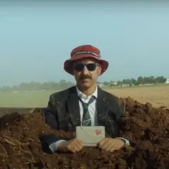 Uomo in abito e cappello rosso, sepolto nel terreno, tiene una lettera con un cuore. Una scena surreale per la recensione "il migliore dei mondi".