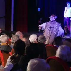Attori in costume sul palco, il pubblico osserva la rappresentazione teatrale, infrangendo la **quarta parete teatro**.