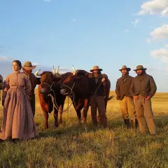 Un gruppo di persone in abiti d'epoca, con due buoi, in un campo dorato. Scena da "La ballata di Buster Scruggs", un film che merita una recensione.