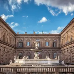 Cortile del Palazzo Pitti con fontana centrale e statue, sotto un cielo azzurro con nuvole bianche.