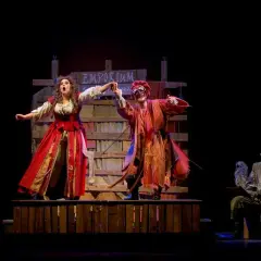 Scena teatrale con due attori in costume, uno con abito rosso e l'altra con maschera e vestiti arancioni. Sullo sfondo un cartello "Emporium". Le recensioni per Romeo è Giulietta lodano queste performance.