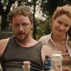 Una coppia sorride, un uomo con barba e canottiera, una donna con capelli rossi. La scena evoca un'atmosfera da "speak no evil recensione".