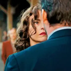 Una donna con labbra rosse e capelli ricci guarda intensamente un uomo, il segreto del suo volto trama. Sullo sfondo, persone sfocate in una stazione.
