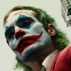 Il volto truccato di chi è Joker, con capelli verdi e un sorriso rosso dipinto, guarda verso l'alto.