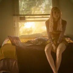 Una ragazza bionda con un vestito a quadri siede sul letto, illuminata dalla luce del tramonto. Una scena che fa pensare a una recensione di Euphoria.