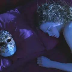 Una donna dorme su un letto di seta viola, accanto a una maschera veneziana ornata. La sua curiosità è palpabile, come se stesse sognando occhi chiusi.