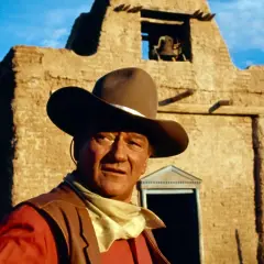 John Wayne, icona del West, con cappello e bandana, davanti a una chiesa diroccata. Ricorda le sue frasi western famose.