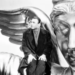 Un uomo seduto su un'ala d'angelo dorata, con un volto statuario di fronte. Un'immagine iconica per capire "il cielo sopra berlino".