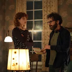 Una donna con capelli rossi e un uomo con occhiali discutono animatamente in una stanza. Lui tiene un quaderno, forse sto pensando di finirla qui recensioni.