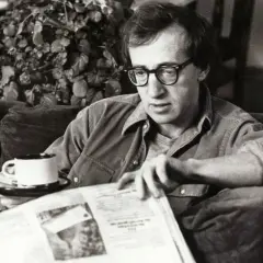 Woody Allen, con occhiali e camicia, legge un giornale con una tazza di caffè, forse pensando al suo prossimo Oscar.