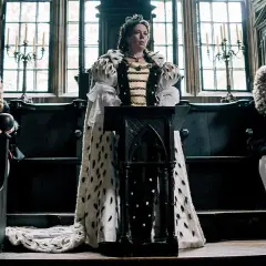 Regina Anna, la favorita, in abiti regali, ascolta attentamente mentre consiglieri con parrucche elaborate la osservano.
