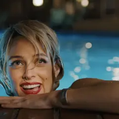 Una donna sorride dall'acqua di una piscina, con luci sfocate sullo sfondo. Una scena che ricorda "Under the Silver Lake".