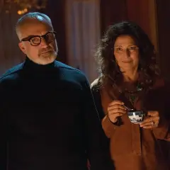 Coppia anziana in un interno buio, lui con occhiali e dolcevita nero, lei sorride con una tazza in mano. Sembra il **get out finale**.