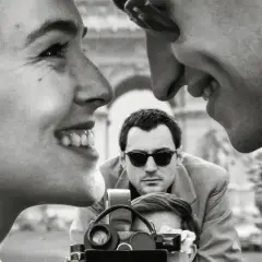 Primo piano di una donna sorridente, con un uomo che la guarda e un altro uomo con occhiali da sole che filma. Un omaggio ai registi importanti.