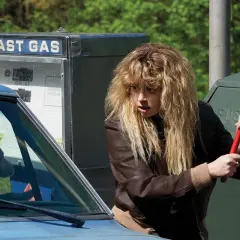 La protagonista di Poker Face, Nadia Vulvokov, con i suoi capelli biondi spettinati, impugna un tergivetro rosso davanti a una pompa di benzina "Last Gas".