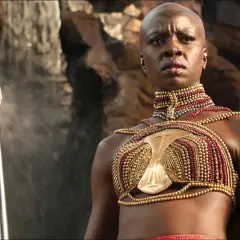 Guerriera Wakandiana con lancia, indossa un elaborato costume dorato e rosso, degno di un premio Oscar ai migliori costumi.