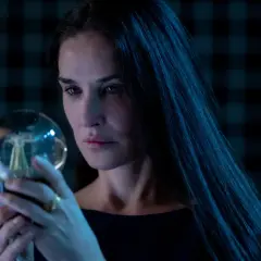 Donna con lunghi capelli scuri osserva una sfera luminosa, un'immagine che evoca mistero e riflessione, come in una recensione di "The Substance".