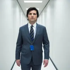 Uomo in abito grigio cammina in un corridoio bianco, con un badge blu appeso al collo. La sua espressione è seria, quasi a suggerire una scissione nella sua recensione.