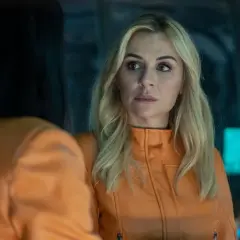 Donna bionda in giacca arancione parla con una persona con capelli scuri. Forse una scena da "il rifugio atomico recensioni".