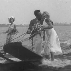 Coppia di sposi baciarsi su una barca trainata, con un uomo in cilindro e una donna con cappello che li seguono. Un'immagine che sembra uscita da un film di 100 anni fa.
