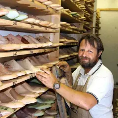 Un calzolaio sorridente in bottega, circondato da forme di legno per scarpe. Sembra Daniel Day Lewis, immerso nel suo mestiere.