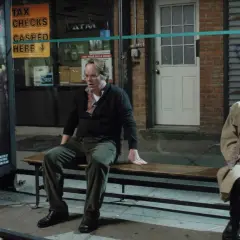 Uomo e donna seduti su una panchina, vicino a un poster di "Little Winky". Una scena che evoca la complessità di "Synecdoche, New York".