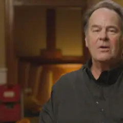 Dan Aykroyd oggi, con un'espressione pensierosa, indossa una camicia scura. Sullo sfondo, oggetti d'arte e sedie rosse.
