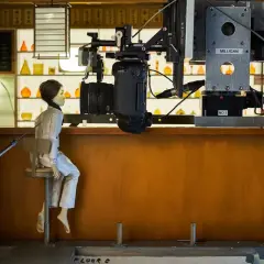 Bambola seduta su uno sgabello, pronta per essere animata in stop motion. La telecamera è posizionata sopra di lei, pronta a catturare ogni movimento.