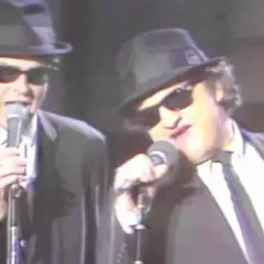Dan Aykroyd giovane e John Belushi sul palco, entrambi con cappello e occhiali da sole, cantano al microfono.