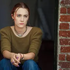 Ragazza pensierosa, con maglione verde e jeans, seduta vicino a un muro di mattoni rossi. Una scena che ricorda la recensione di Lady Bird.