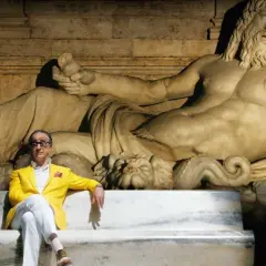Un uomo in giacca gialla seduto su una panchina di marmo, con una statua antica alle spalle. Un'immagine che evoca la grande bellezza, spiegazione visiva dell'arte e del tempo.