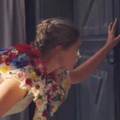 Una ragazza con un vestito fiorito, capelli raccolti, spinge una porta scura. Un'immagine che evoca la recensione di Midsommar.