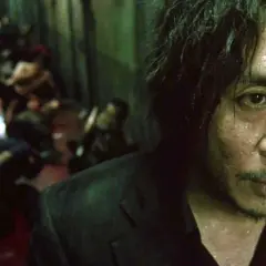 Primo piano di un uomo sudato, con un'espressione intensa, sullo sfondo una scena confusa di persone. Ricorda il film Oldboy e il suo polpo.