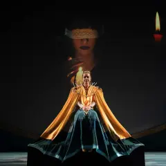 Scena teatrale ispirata al circe film: una donna con mantello dorato, circondata da candele, sotto un'immagine proiettata di una figura bendata.