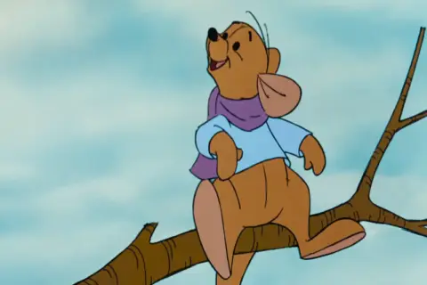 Winnie the Pooh, interpretato da Jim Cummings, siede su un ramo con una sciarpa viola e una camicia azzurra.
