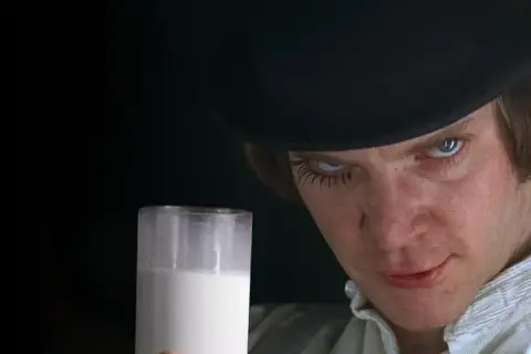 Un uomo con un cappello nero e ciglia finte tiene un bicchiere di latte, rompendo la quarta parete con uno sguardo intenso.