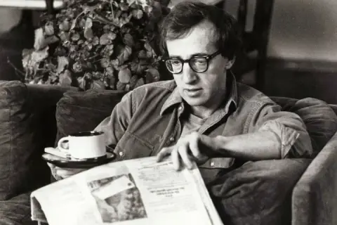 Woody Allen, con occhiali e camicia, legge un giornale con una tazza di caffè, forse pensando al suo prossimo Oscar.