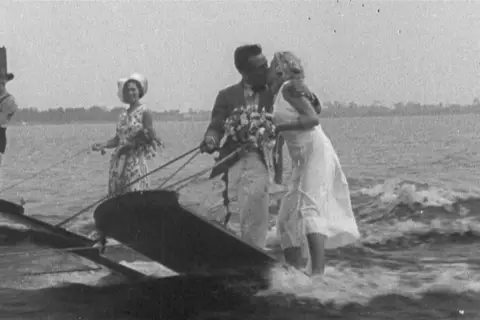 Coppia di sposi baciarsi su una barca trainata, con un uomo in cilindro e una donna con cappello che li seguono. Un'immagine che sembra uscita da un film di 100 anni fa.