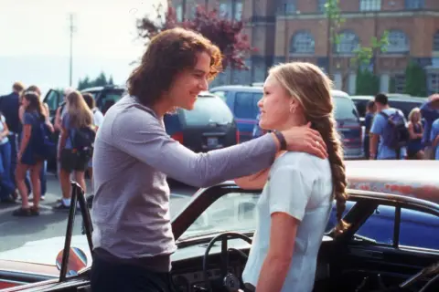 Il cast di "10 cose che odio di te" con Heath Ledger e Julia Stiles che si guardano sorridendo vicino a un'auto.