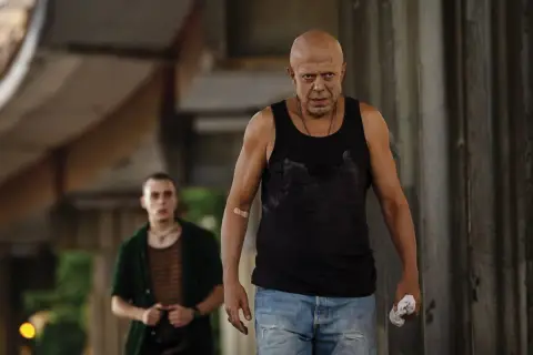 Due uomini, uno calvo e con un cerotto sul braccio, camminano in un ambiente urbano. Scena dal film "Adagio".