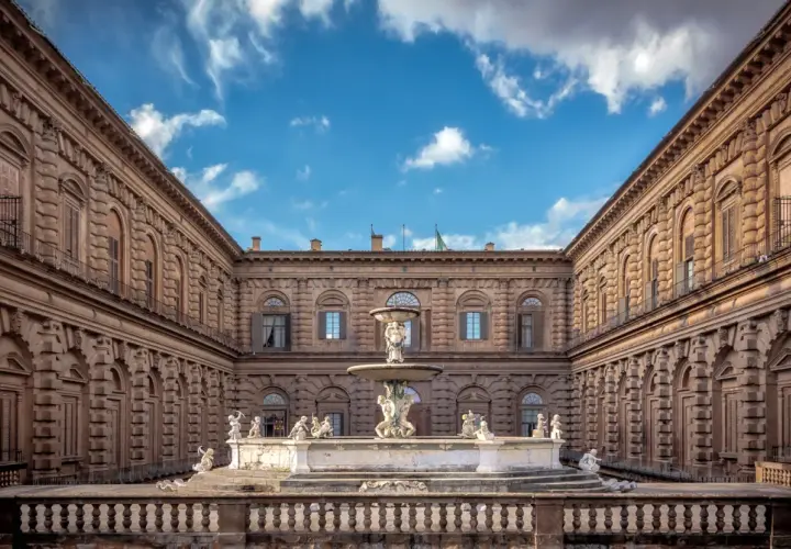 Cortile del Palazzo Pitti con fontana centrale e statue, sotto un cielo azzurro con nuvole bianche.