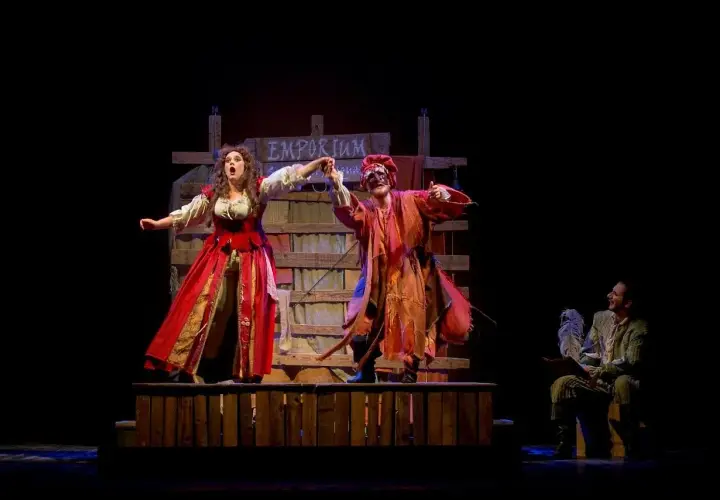 Scena teatrale con due attori in costume, uno con abito rosso e l'altra con maschera e vestiti arancioni. Sullo sfondo un cartello "Emporium". Le recensioni per Romeo è Giulietta lodano queste performance.