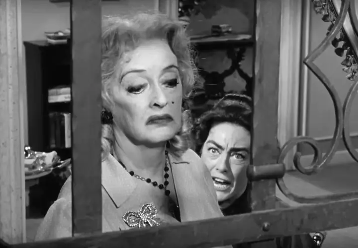 Bette Davis anziana, con un'espressione di disgusto, è dietro una grata. Un'altra donna con occhi sbarrati la spia.