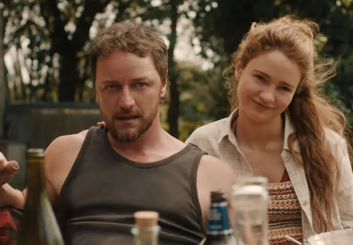 Una coppia sorride, un uomo con barba e canottiera, una donna con capelli rossi. La scena evoca un'atmosfera da "speak no evil recensione".