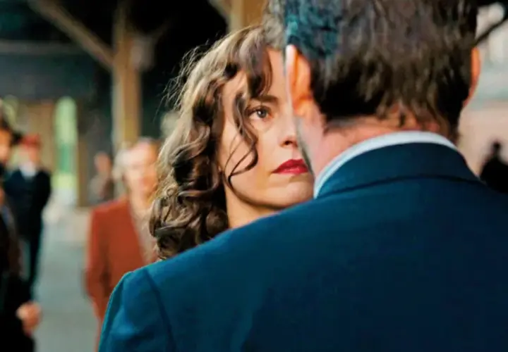 Una donna con labbra rosse e capelli ricci guarda intensamente un uomo, il segreto del suo volto trama. Sullo sfondo, persone sfocate in una stazione.